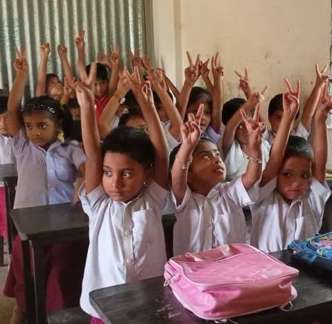 Children at Monobikash School raising hands with peace signs in class